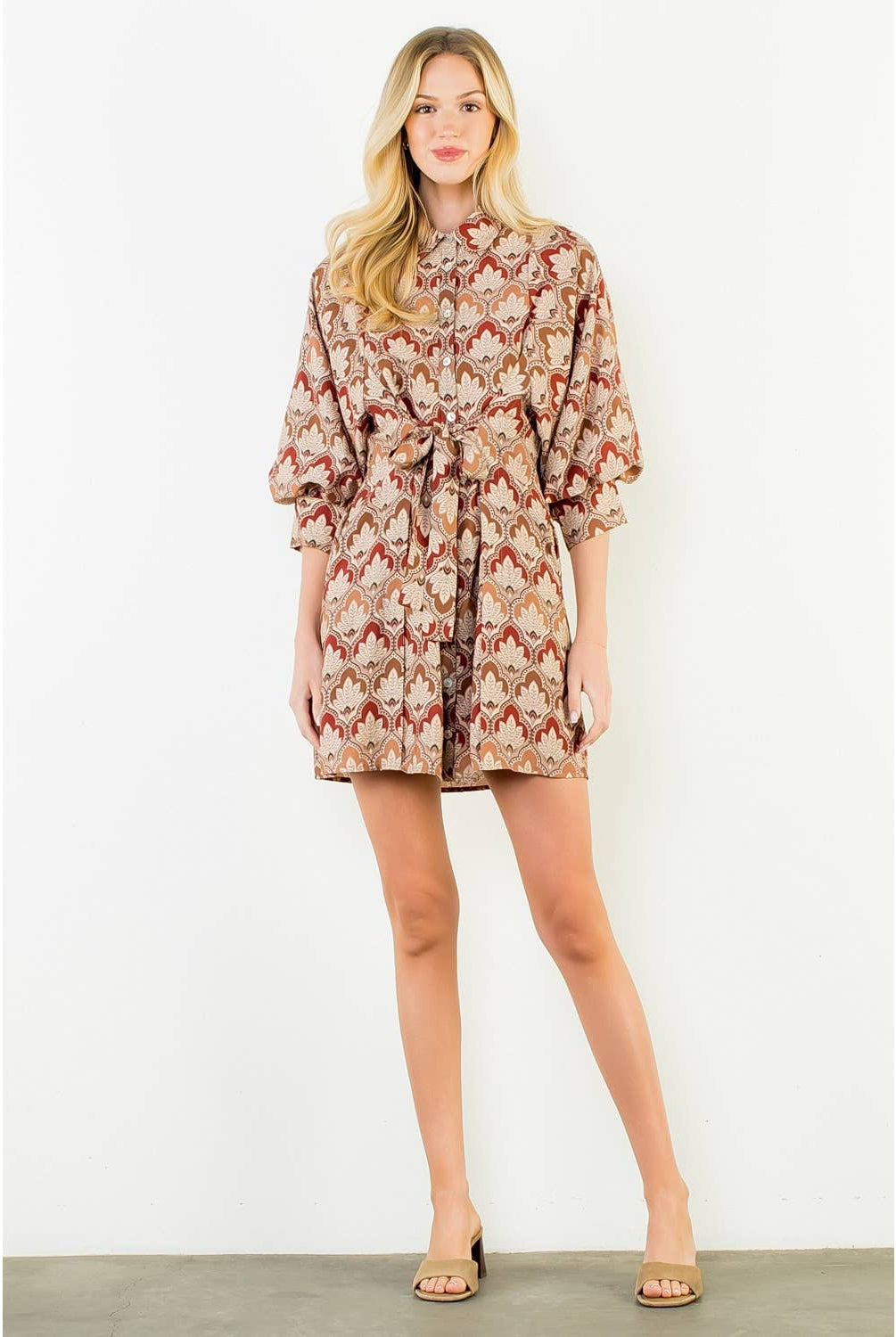 A woman modeling a short wrap-style dress with a beige, brown, and red floral pattern, featuring three-quarter puffed sleeves and a self-tie belt, set against a plain white studio background.