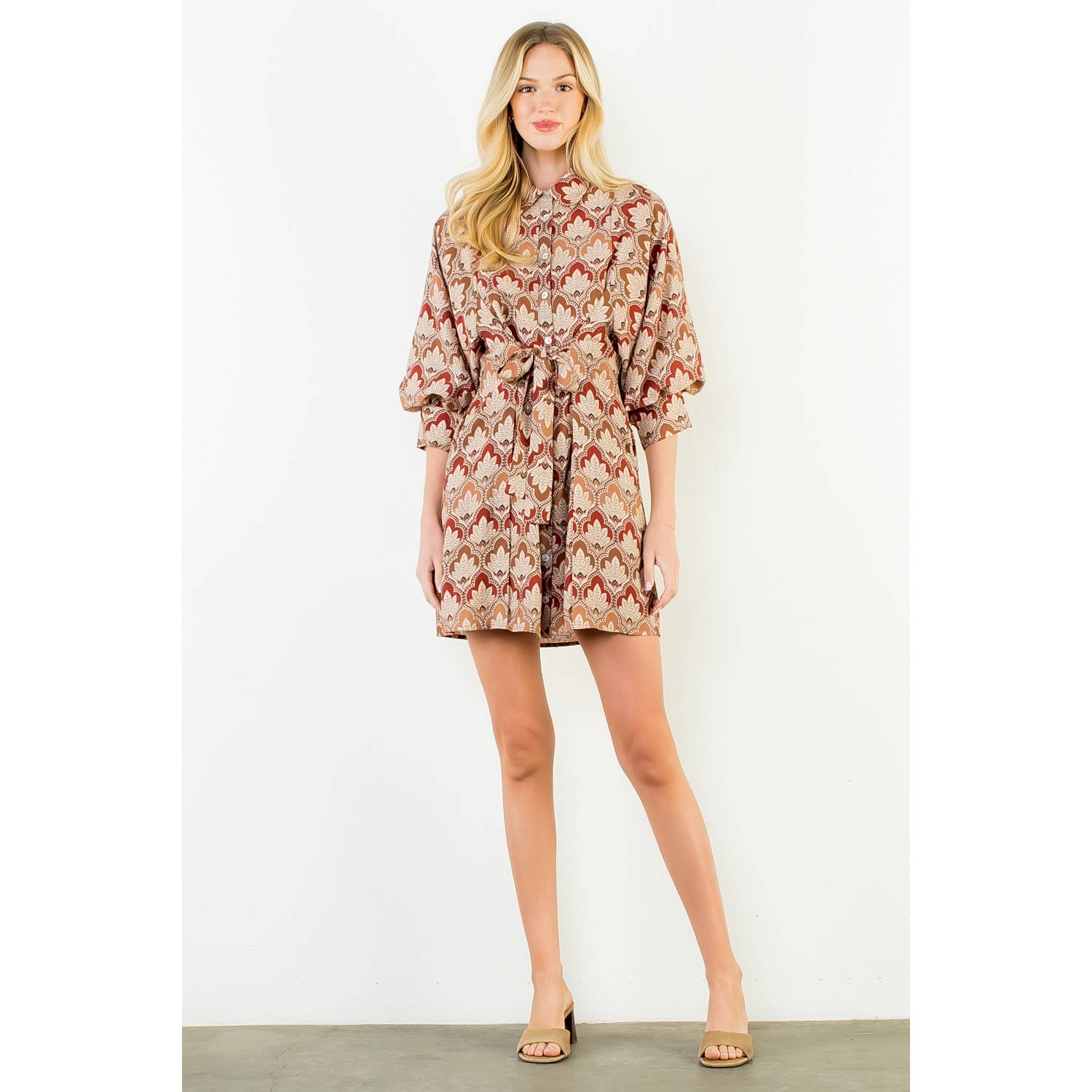 A woman modeling a short wrap-style dress with a beige, brown, and red floral pattern, featuring three-quarter puffed sleeves and a self-tie belt, set against a plain white studio background.