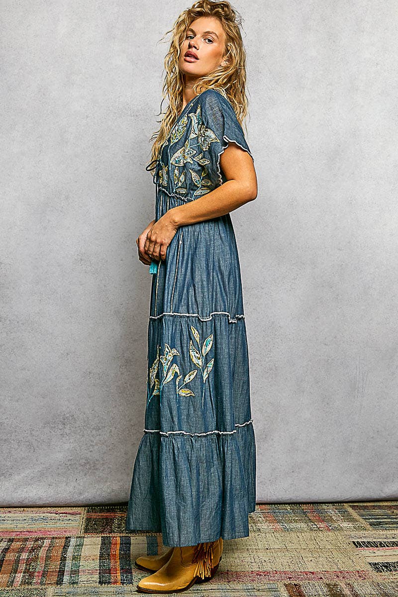 Woman wearing a long denim dress with floral embroidery against a gray wall.