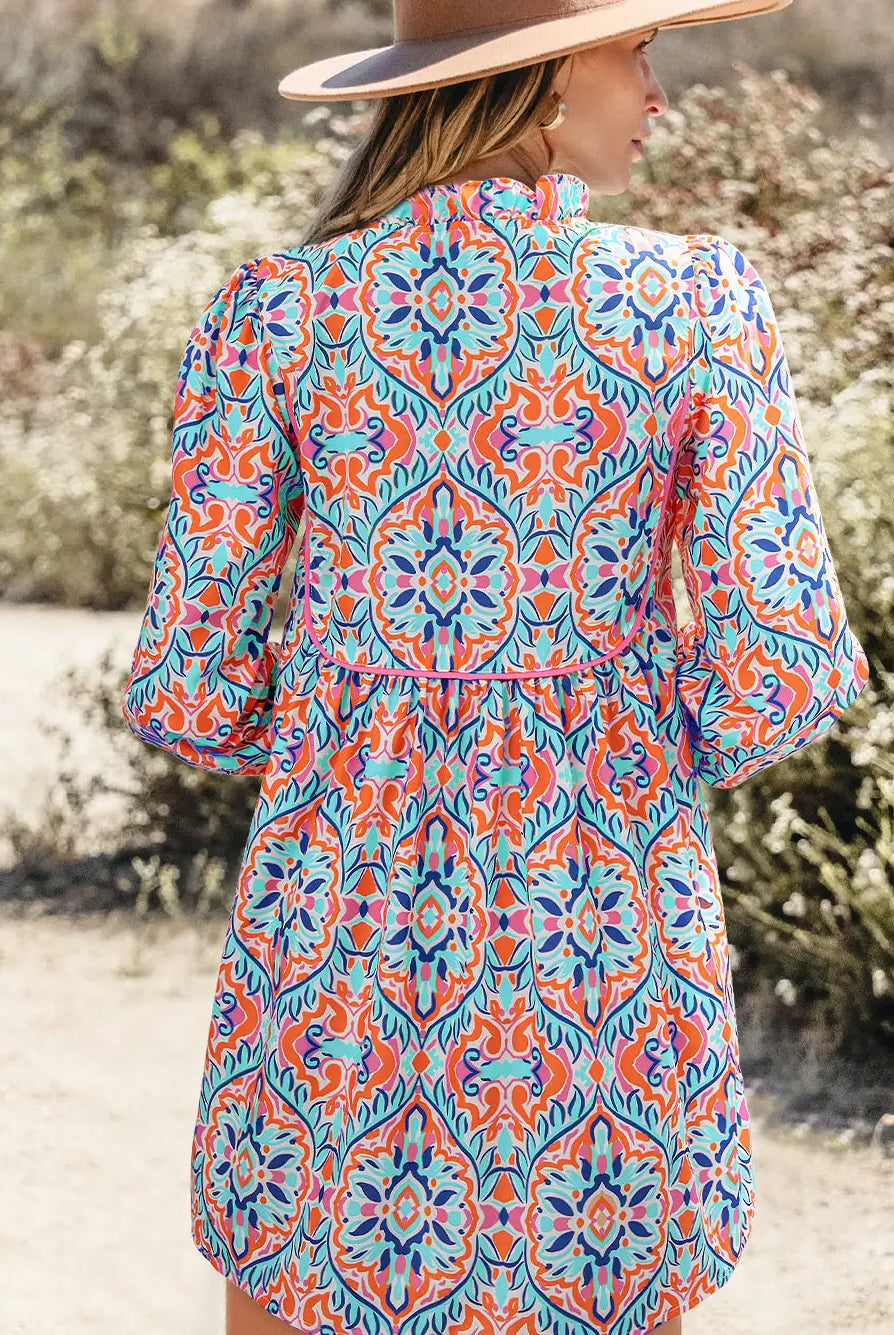 A woman wearing a colorful bohemian-style patterned tunic dress and a wide-brimmed hat, photographed from behind in an outdoor desert setting.
