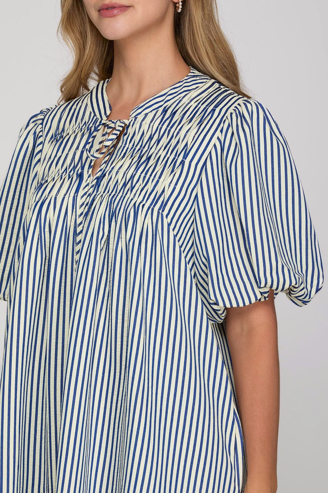 A close-up of a woman wearing a blue and white vertically striped blouse with puff sleeves and a smocked neckline.
