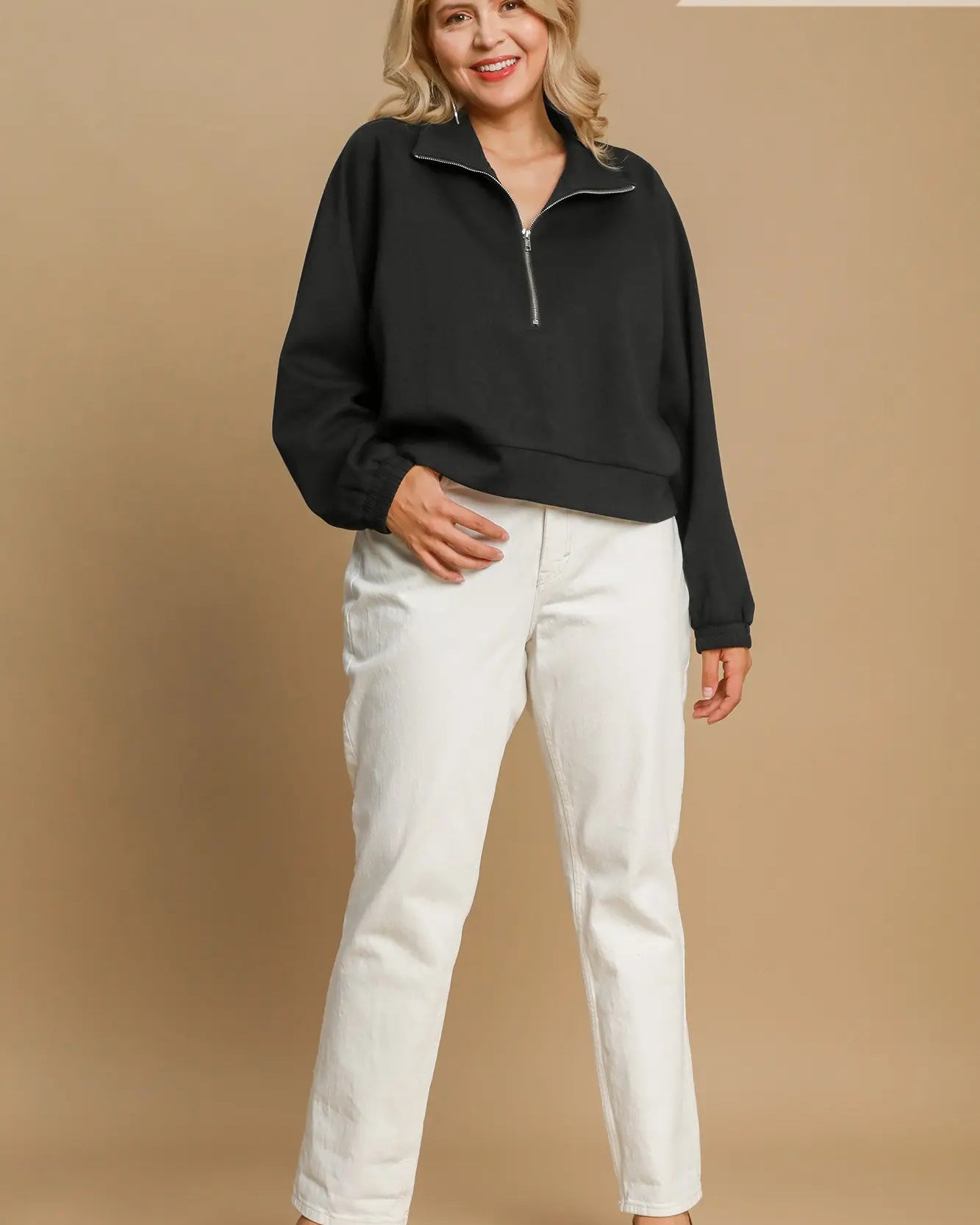 A woman modeling a black long-sleeve quarter-zip sweatshirt with white pants, standing against a solid beige studio background. Text overlays read 'PLUS SIZE' at the top right and 'BLACK' at the bottom right.