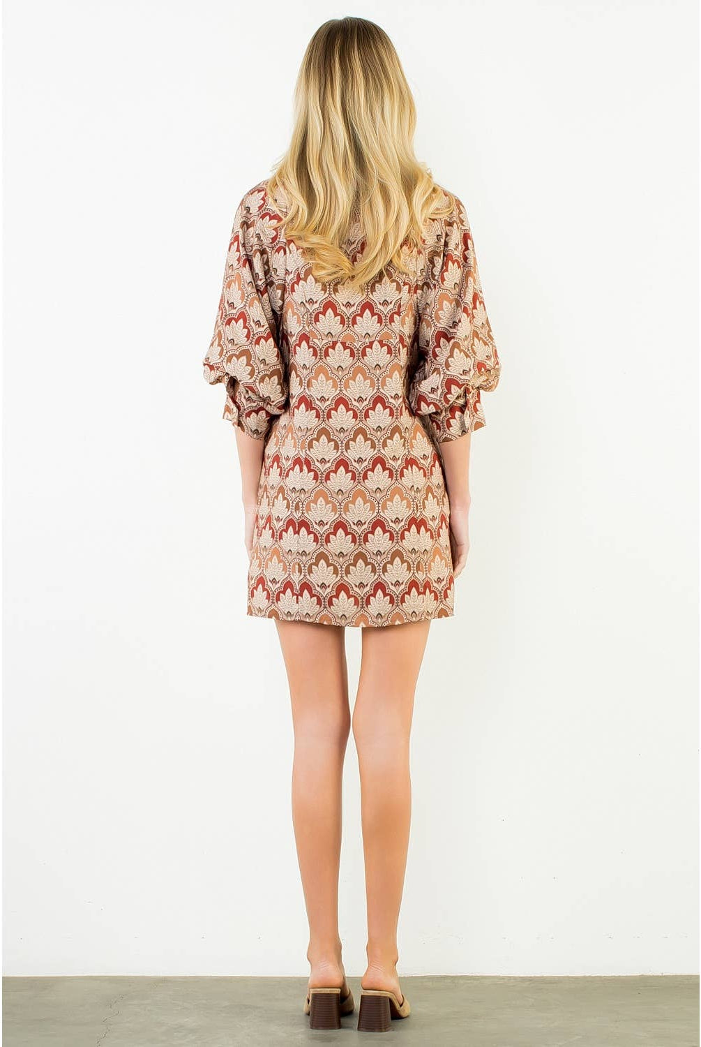 A woman viewed from behind wearing a short puff-sleeve dress with a beige, brown, and rust-red geometric pattern, standing against a plain white wall.