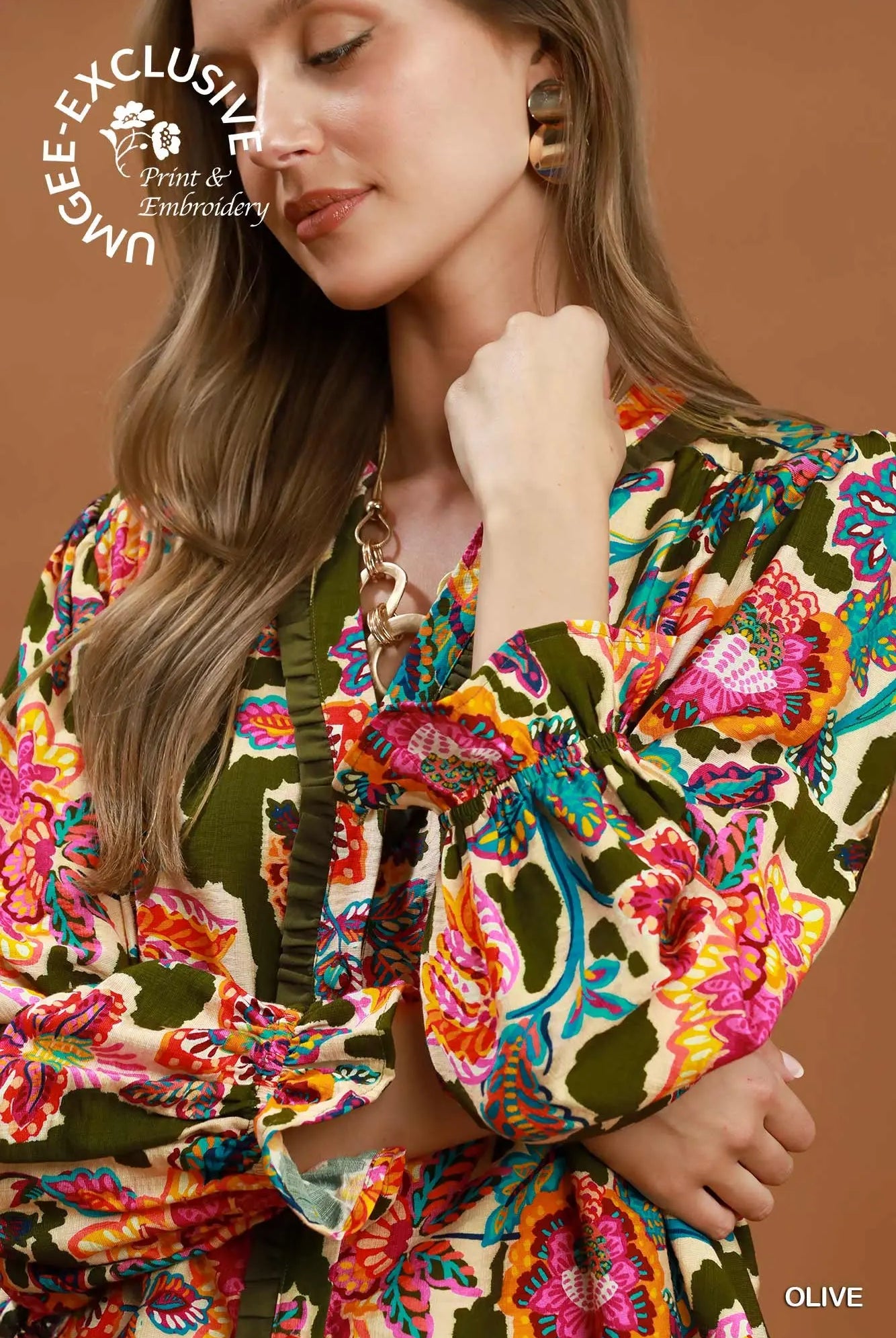 A close-up of a woman wearing a vibrant, multicolored floral print blouse with olive green ruffled trim and puff sleeves, set against a solid warm brown background.