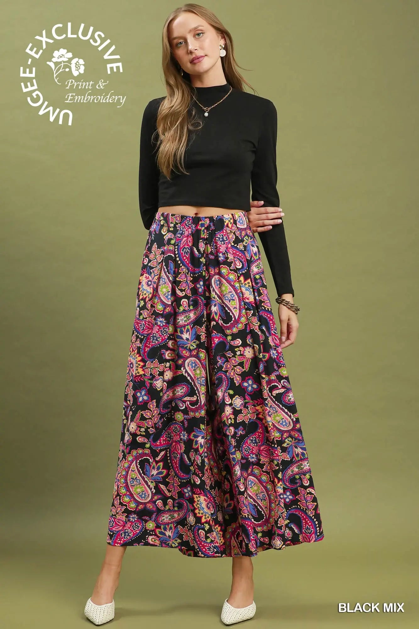 A woman modeling a black long-sleeve crop top and a high-waisted, wide-leg midi skirt with a vibrant paisley print in pink, purple, blue, and yellow on a black base. She is standing against a solid olive green studio background.