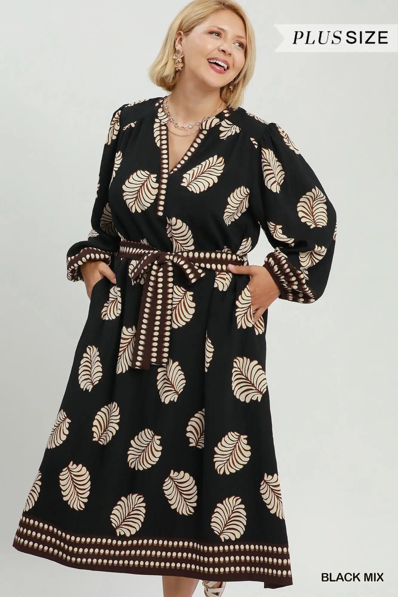 A woman modeling a black long-sleeve maxi dress with a beige palm leaf pattern and a matching tie belt, shown against a light gray studio background with 'PLUS SIZE' text in the top right.