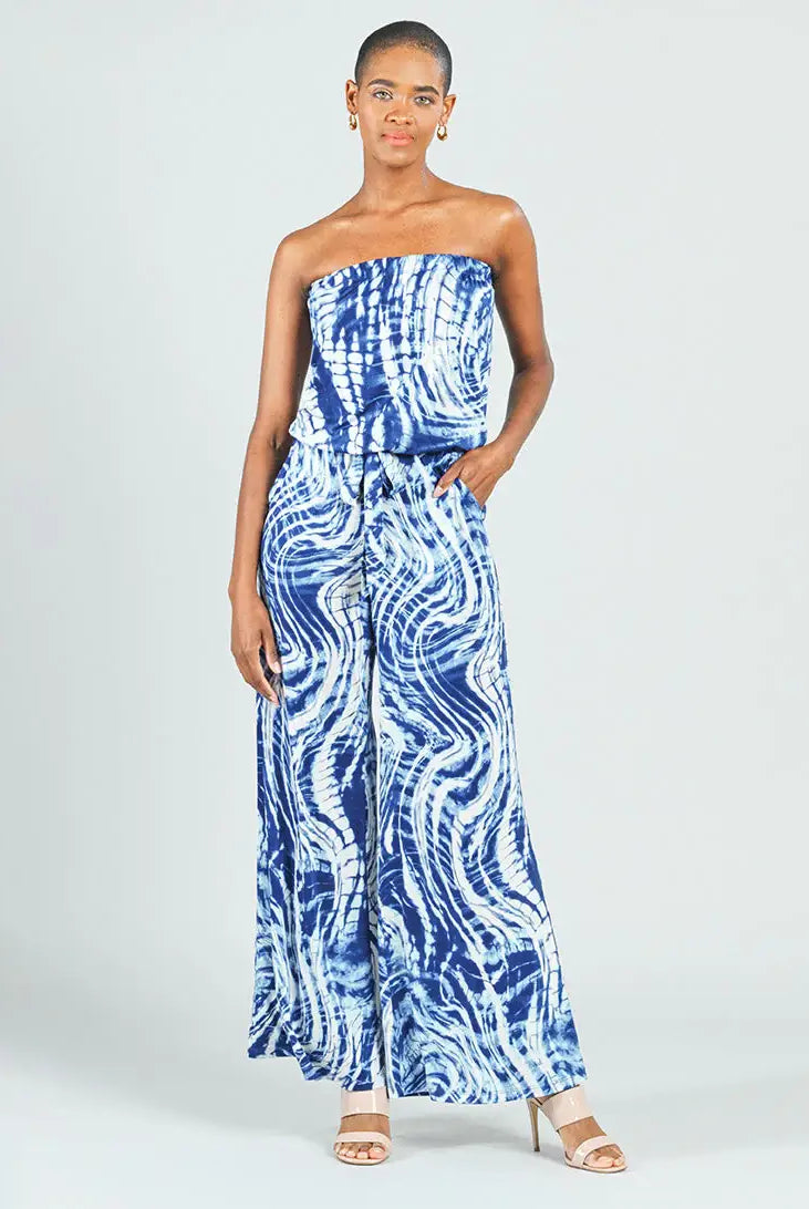 A woman modeling a strapless blue and white tie-dye wide-leg jumpsuit against a light gray studio background.