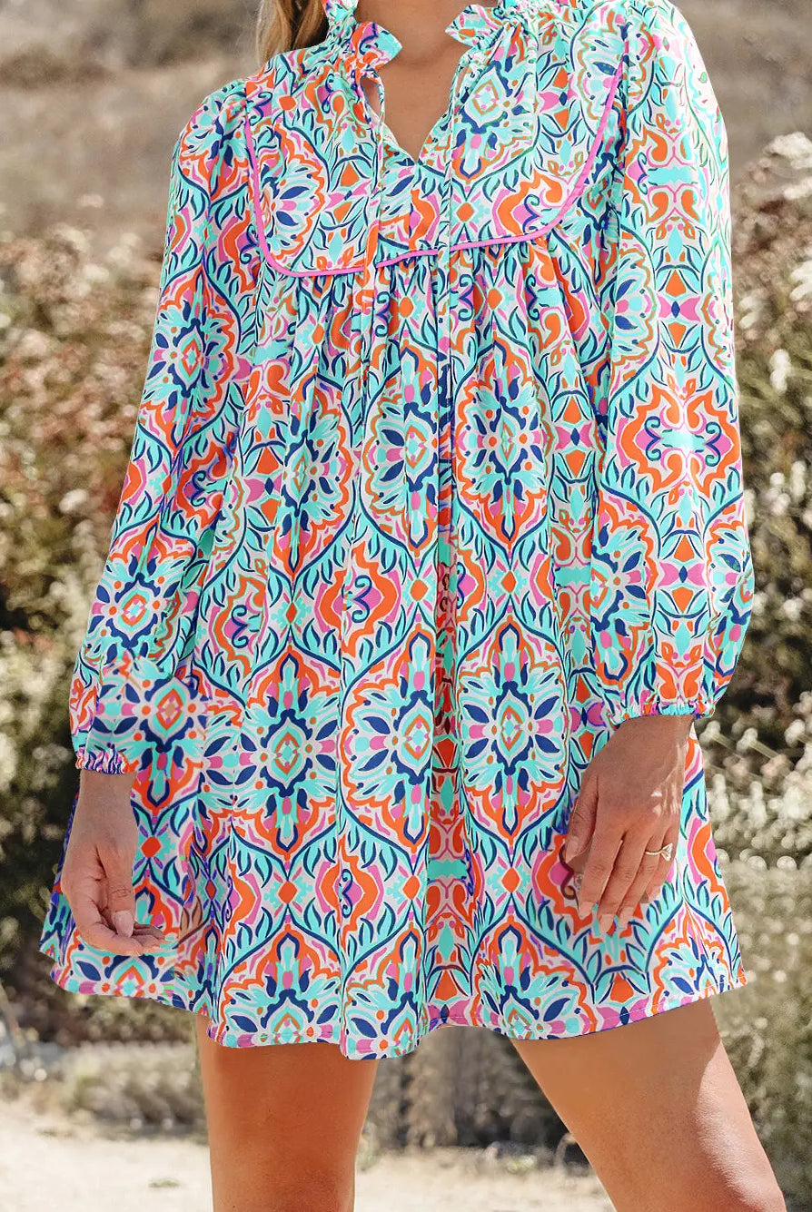 A woman wearing a long-sleeve bohemian-style mini dress with a vibrant multicolor paisley and floral pattern in turquoise, orange, pink, and purple, set against a blurred outdoor background.