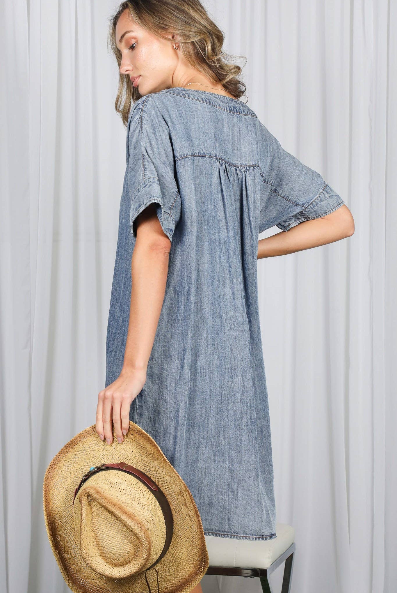Woman wearing a blue dress holding a straw hat against a white curtain background