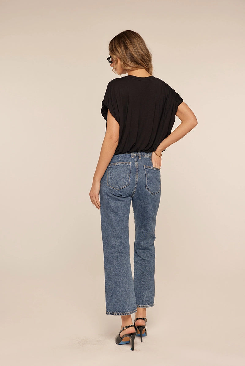A woman is shown from behind wearing blue cropped jeans, a black short-sleeve top, and black high-heeled sandals against a plain beige studio background.