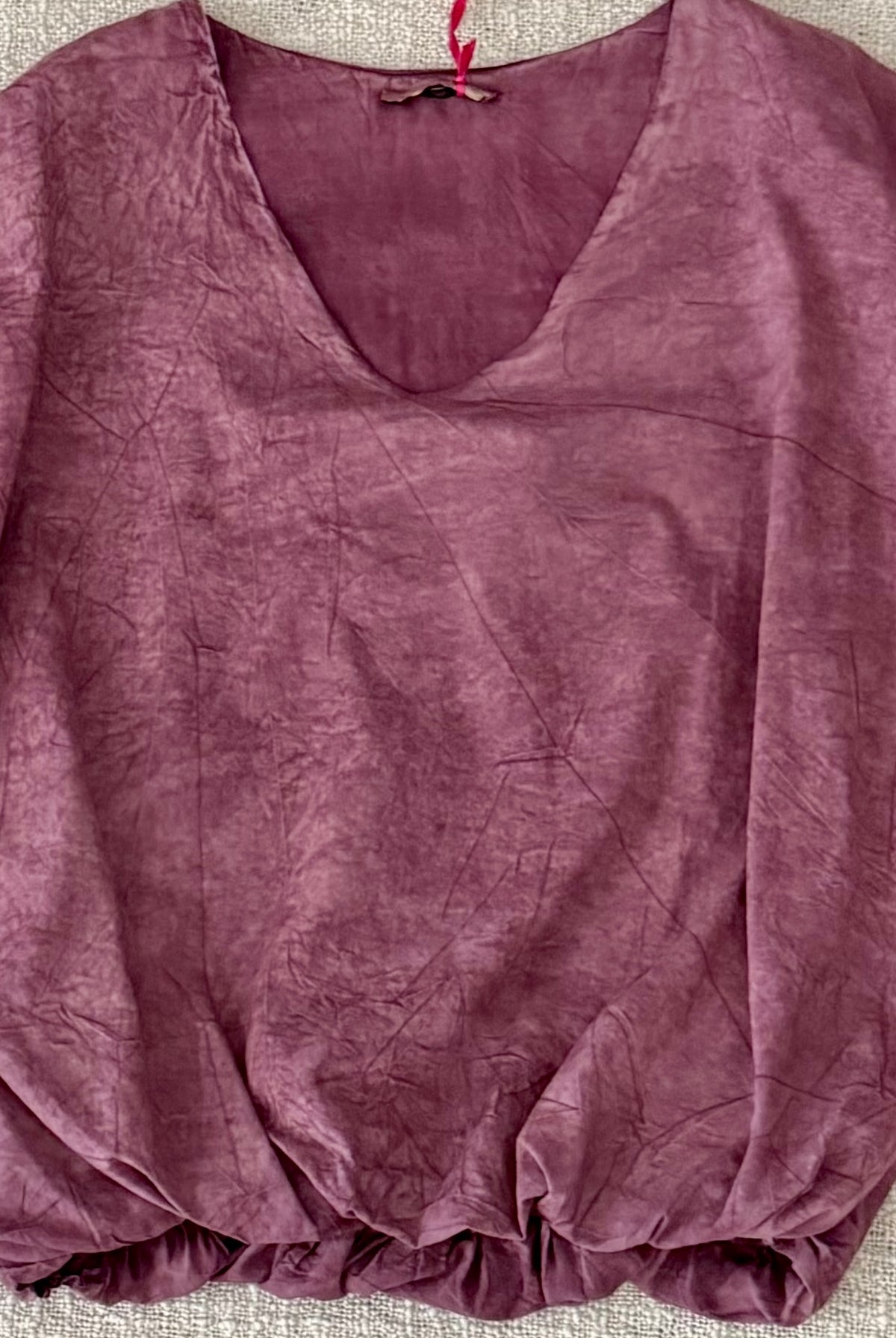 A dusty mauve-purple crinkled cotton blouse with a V-neckline and voluminous elbow-length puff sleeves, laid flat on a textured beige fabric background.