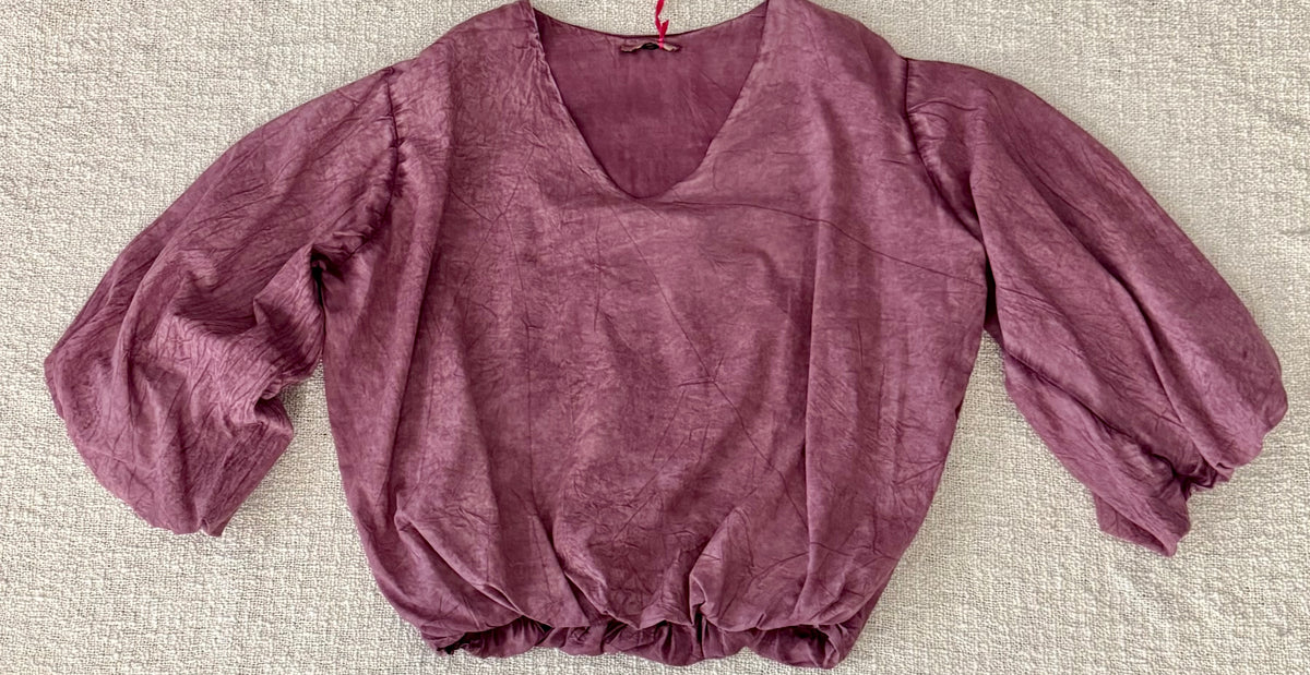 A dusty mauve-purple crinkled cotton blouse with a V-neckline and voluminous elbow-length puff sleeves, laid flat on a textured beige fabric background.