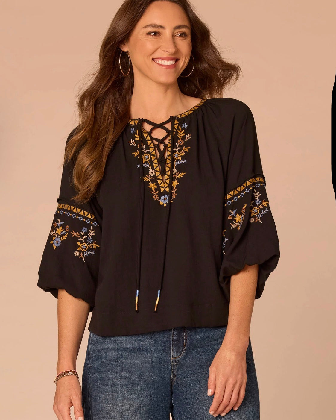 A woman wearing a black bohemian-style blouse with gold, blue, and white floral embroidery on the neckline and sleeves, paired with blue jeans, set against a solid beige background.