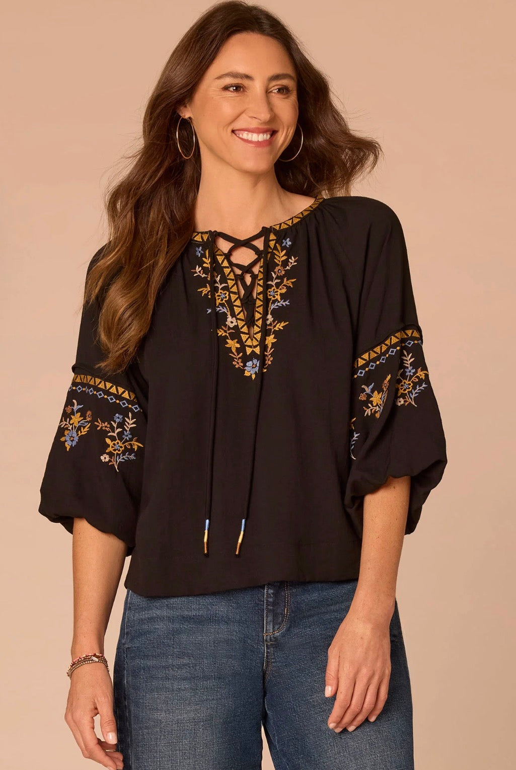 A woman wearing a black bohemian-style blouse with gold, blue, and white floral embroidery on the neckline and sleeves, paired with blue jeans, set against a solid beige background.