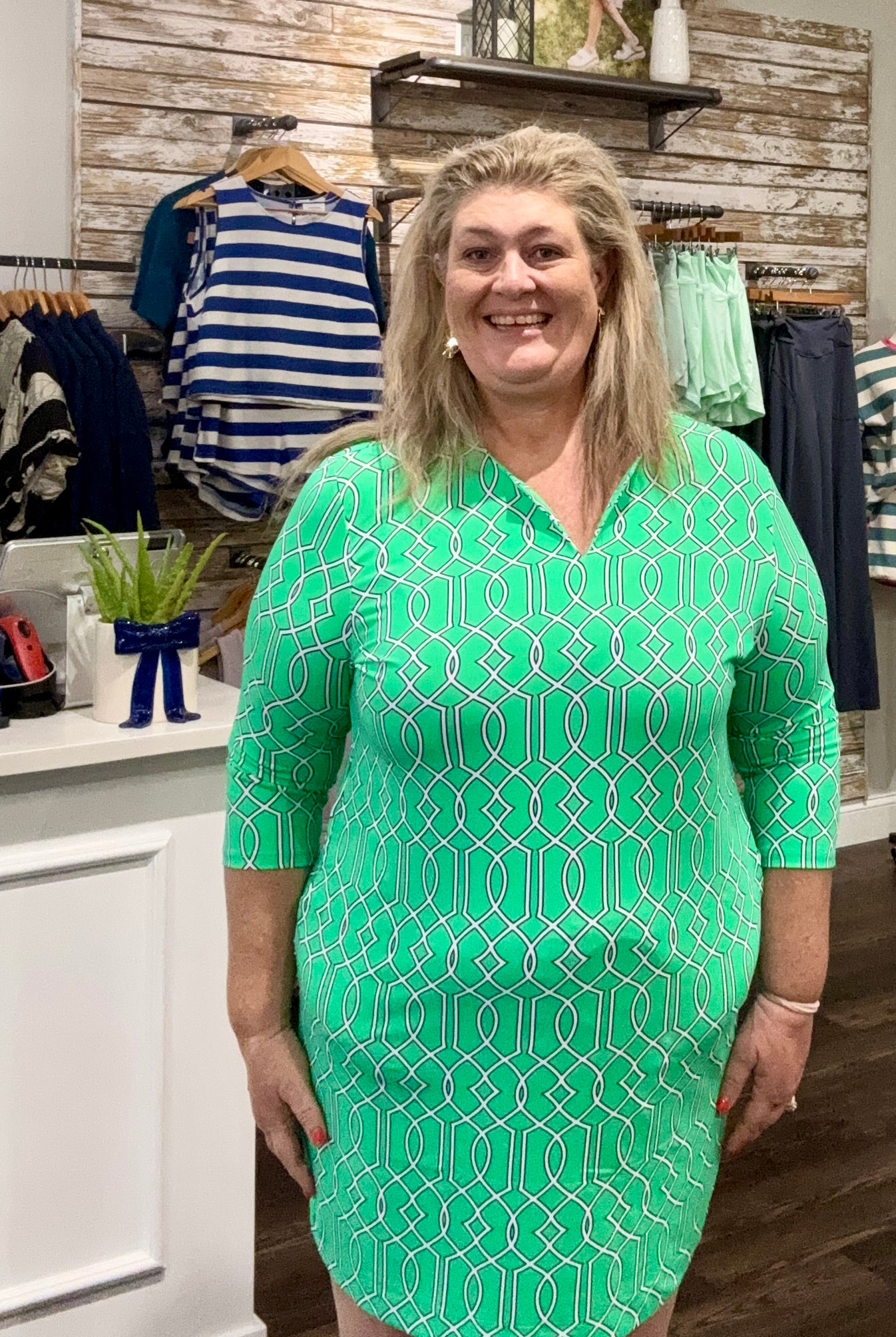 A woman modeling a mint green V-neck dress with a white geometric pattern, standing inside a clothing boutique.