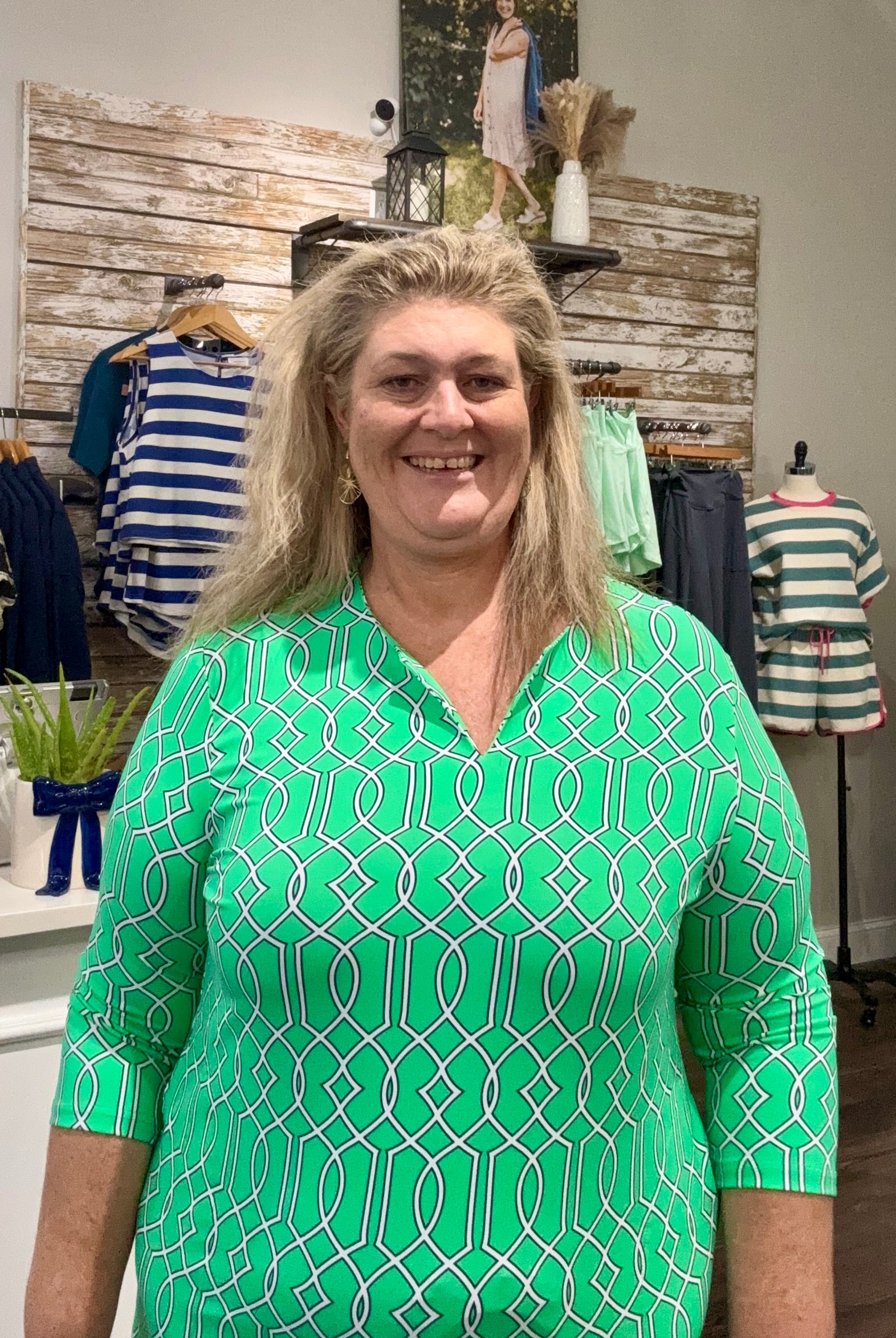 A smiling woman with blonde hair wearing a bright green and white geometric patterned short-sleeve top, standing inside a clothing boutique.