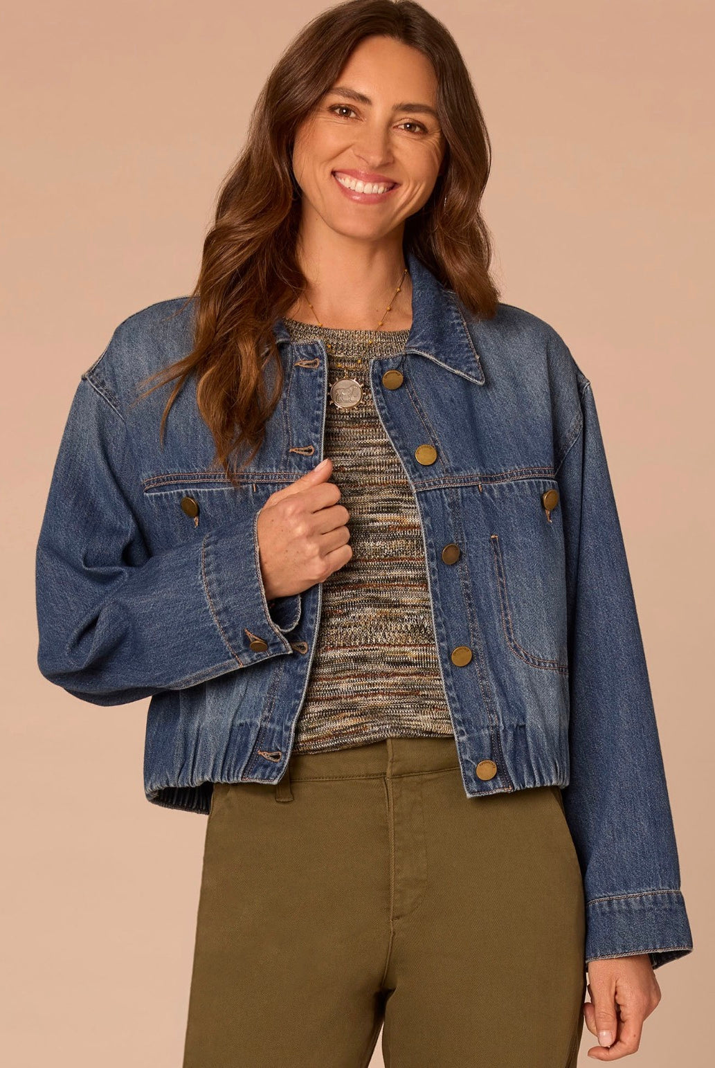 A woman wearing a cropped blue denim jacket over a textured knit top and olive green pants, standing against a solid beige background.