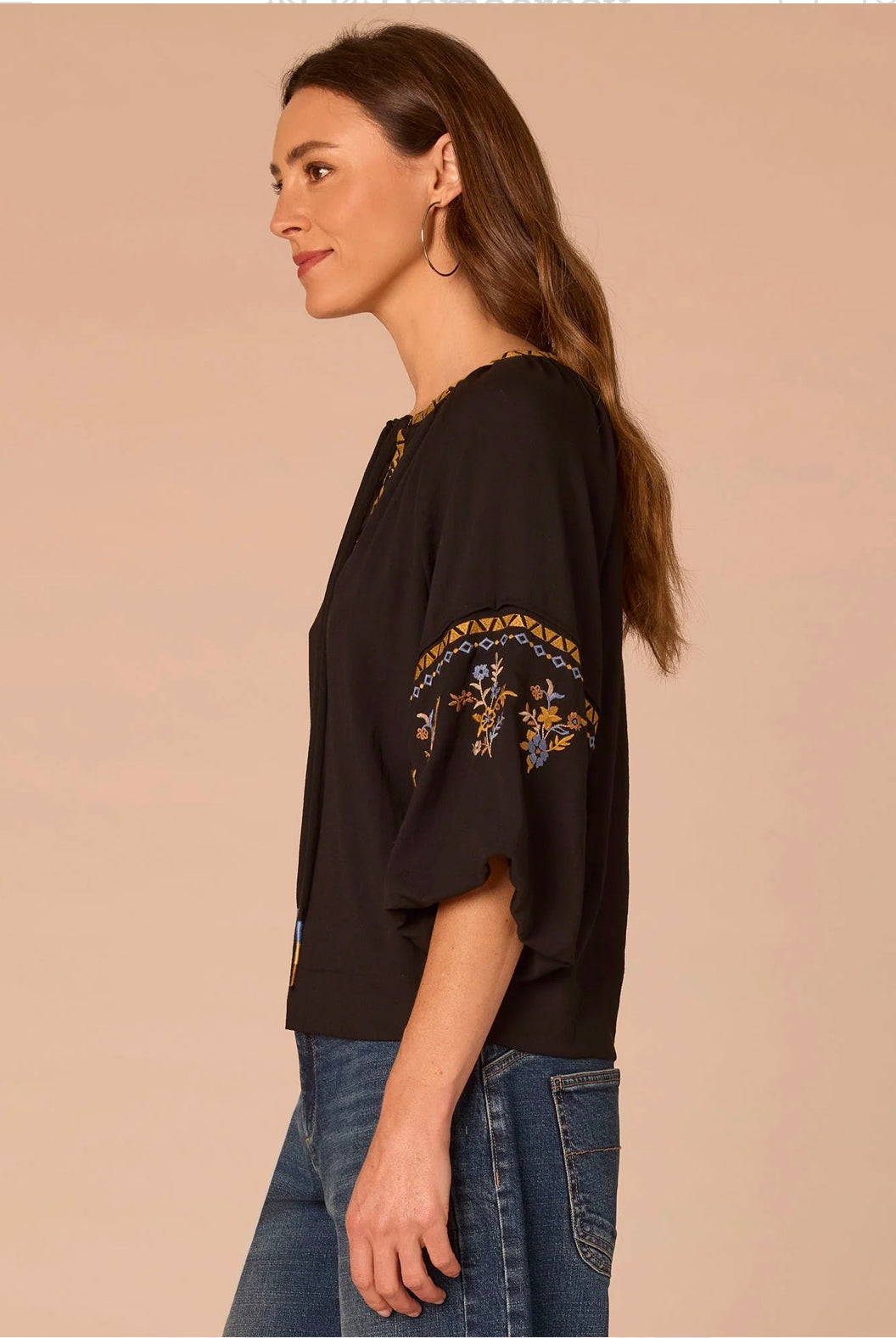 A woman shown in side profile wearing a black short-sleeve top with gold and blue embroidery on the sleeve, paired with blue jeans, against a solid beige background.