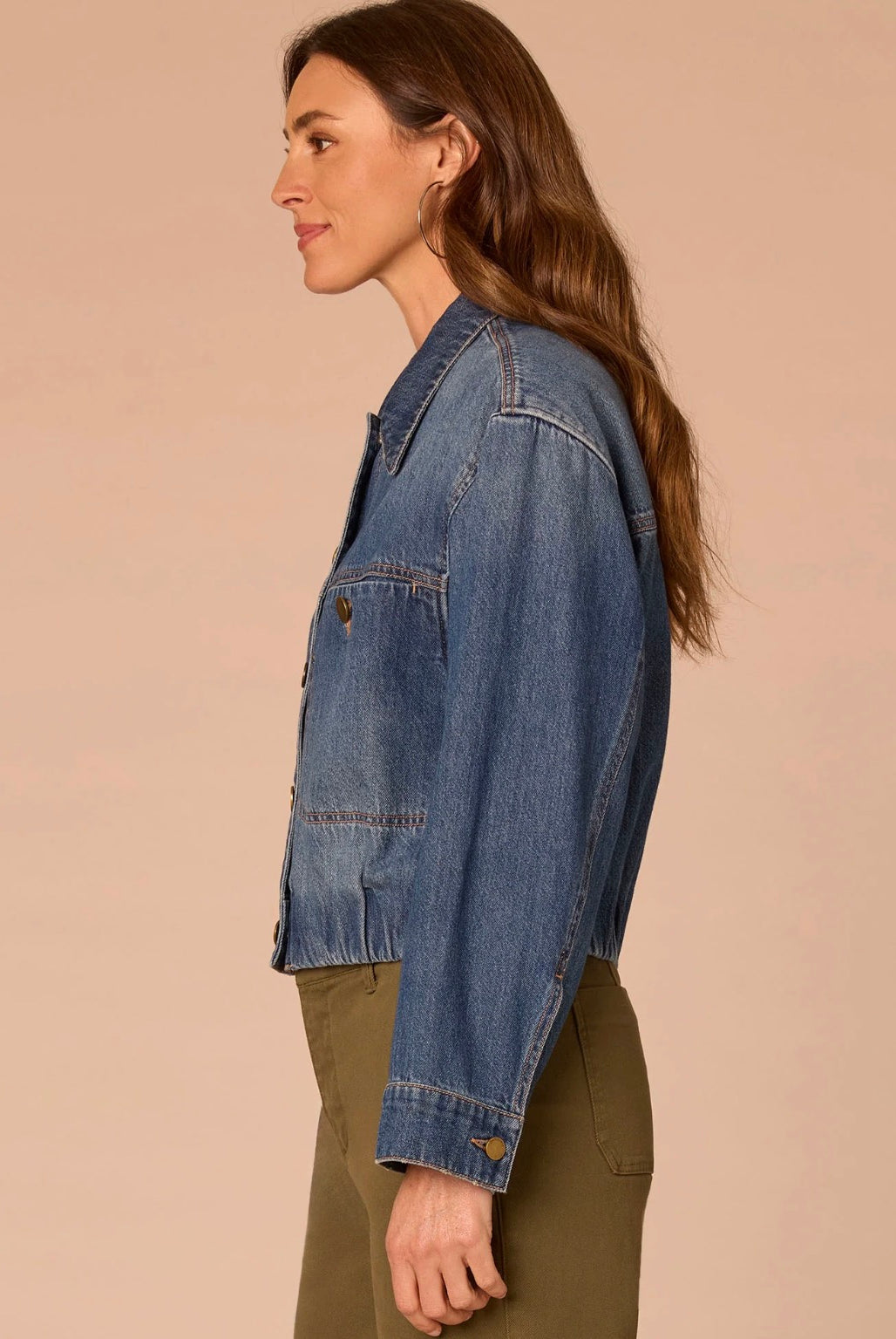 A woman shown in side profile wearing a cropped blue denim jacket over an olive green top, set against a solid warm beige studio background.
