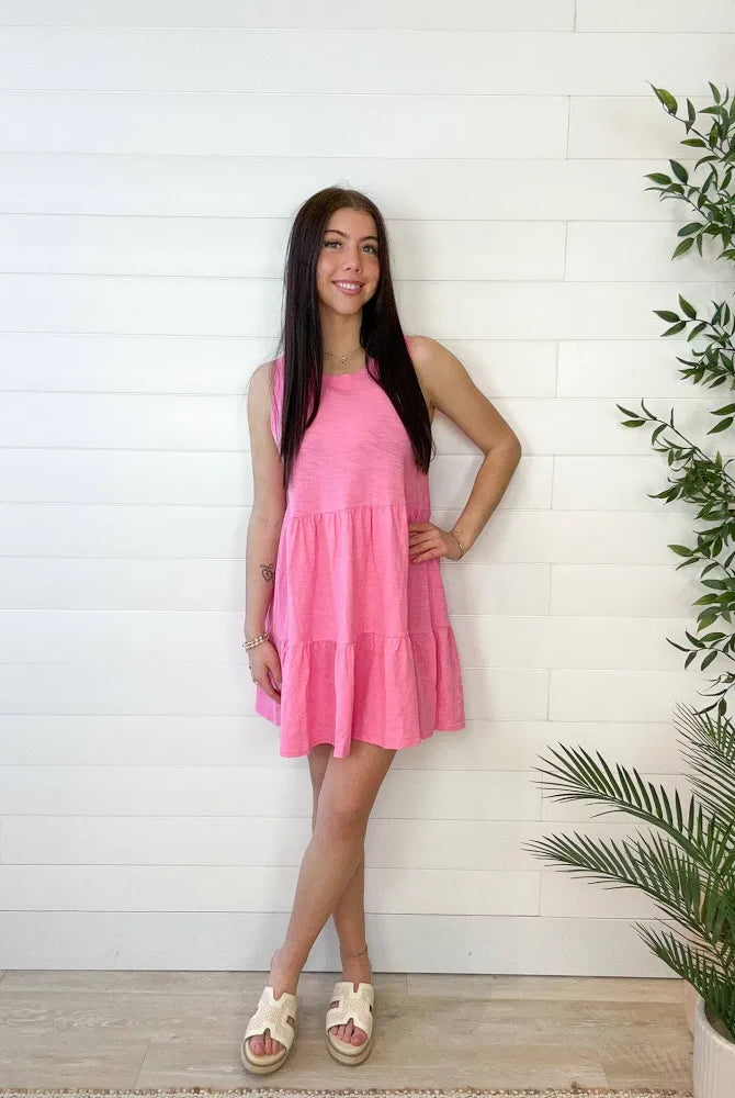 Model wearing tiered, candy, pink, mini dress with neutral wedges against a white wall
