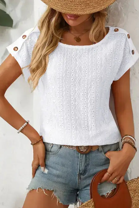 A woman wearing a white short-sleeve eyelet blouse with decorative buttons on the shoulders, paired with denim shorts and a straw hat.