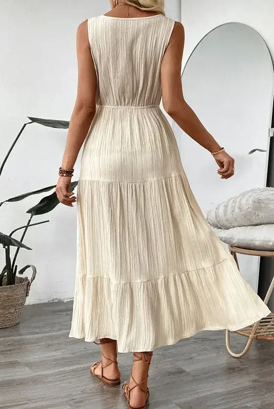Woman wearing a beige pleated dress in a room with a plant and mirror.