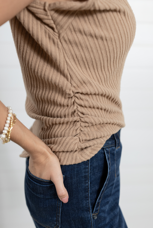 A close-up of a person wearing a beige ribbed knit top with ruching and dark blue jeans, accessorized with pearl and gold bracelets, against a light background.