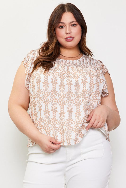 A plus-size woman wearing a short-sleeve top with a beige and white botanical damask pattern, paired with white pants, against a plain white studio background.