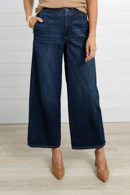 A model wearing dark blue wide-leg cropped jeans, paired with a beige ribbed top and tan pointed-toe heels, standing against a white shiplap wall and light wood floor.