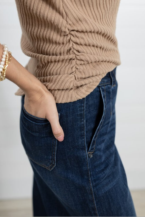 A close-up of a person wearing a beige ruched ribbed knit top and dark blue denim jeans, with one hand in a pocket and pearl-and-gold bracelets on the wrist.