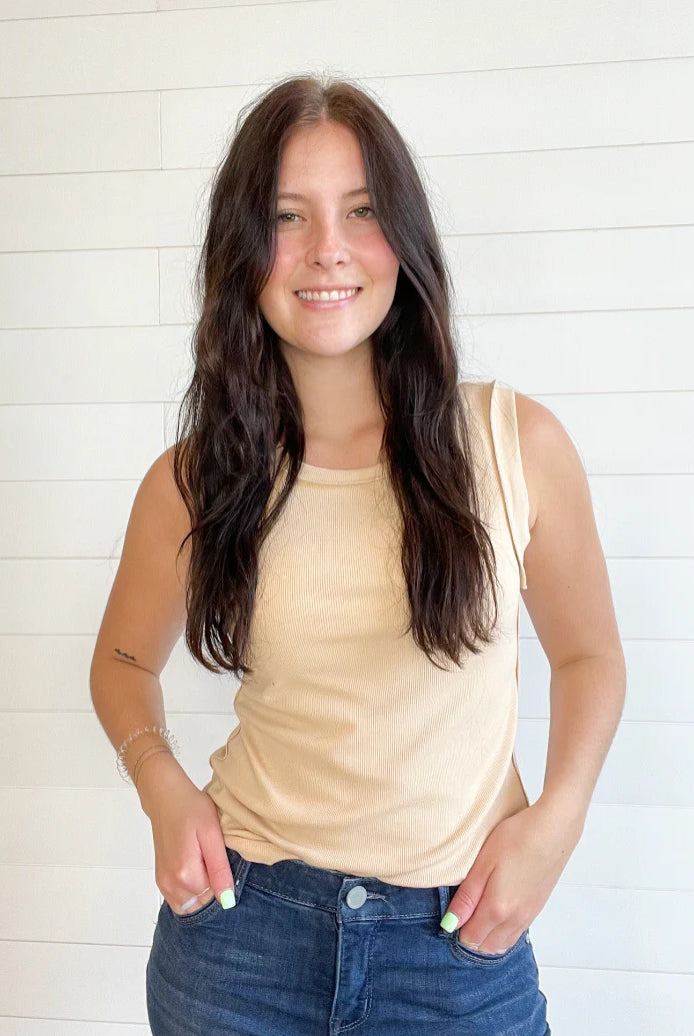 A woman with long dark brown hair smiling, wearing a light beige sleeveless tank top and blue jeans, standing against a white shiplap wall.