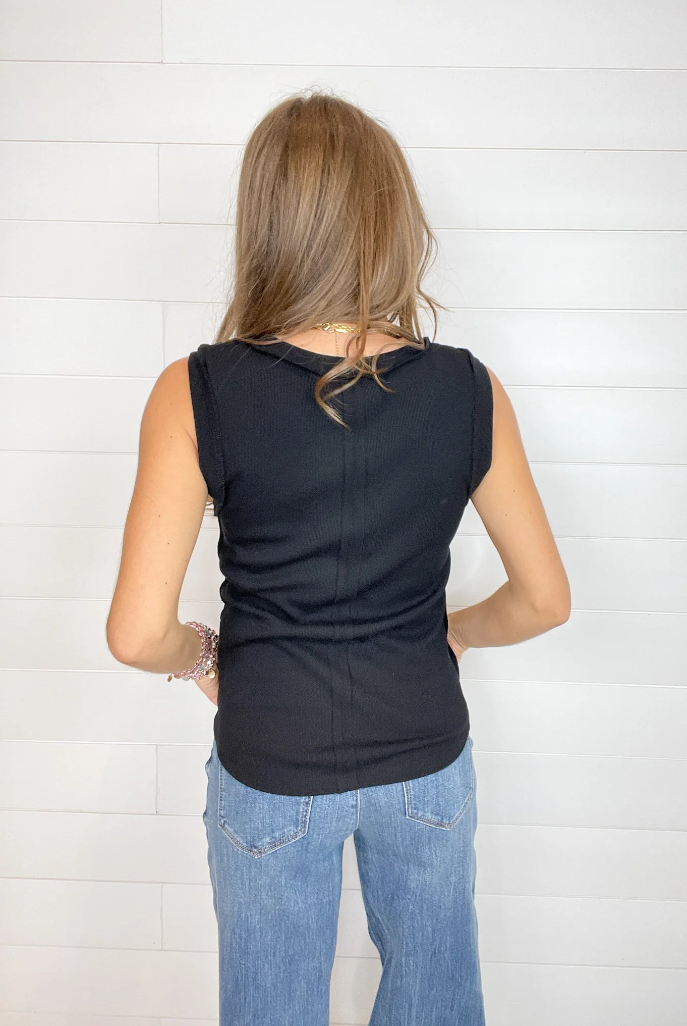A woman viewed from behind wearing a black sleeveless top and light blue jeans, standing against a white shiplap wall.
