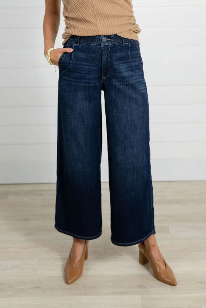 Person wearing blue wide-leg jeans and brown shoes on a light wooden floor.