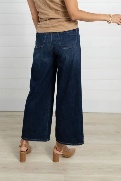 A person shown from the back wearing dark blue wide-leg denim jeans, a beige ribbed knit top, and tan block-heel sandals against a white paneled wall and light wood floor.