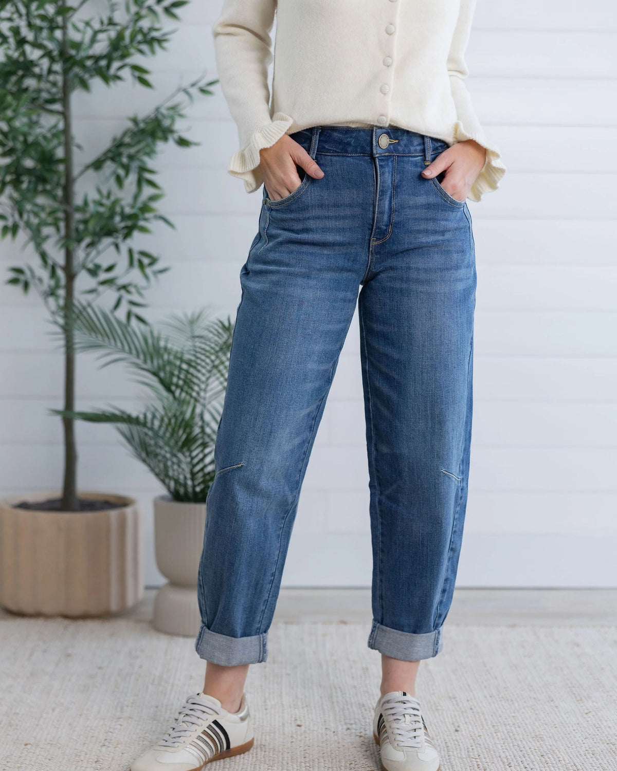 A person wearing blue cuffed jeans, a cream button-up sweater, and white sneakers, standing in front of a white paneled wall with potted plants.