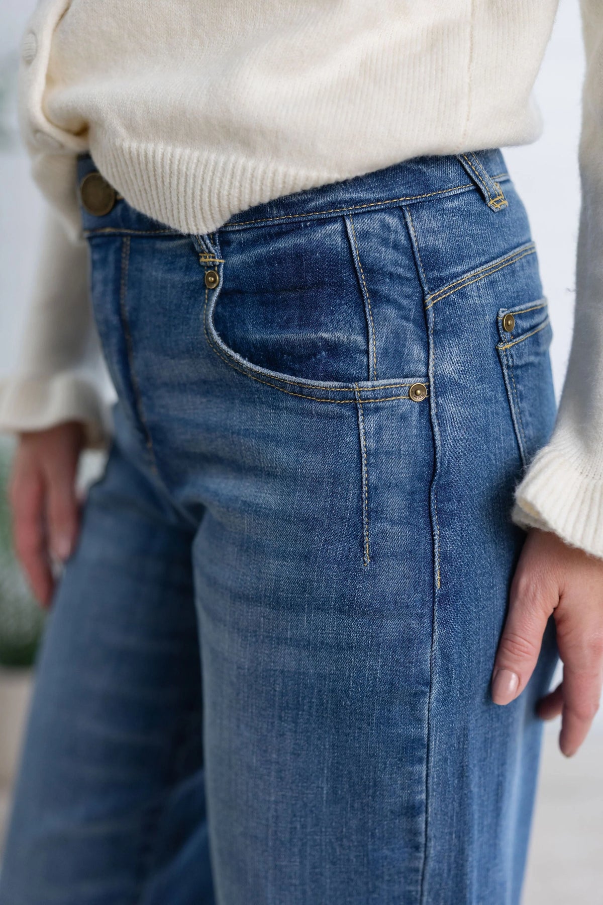 Close-up of a person wearing blue denim jeans with a cream ribbed knit sweater, focusing on the back pocket and waistband details.