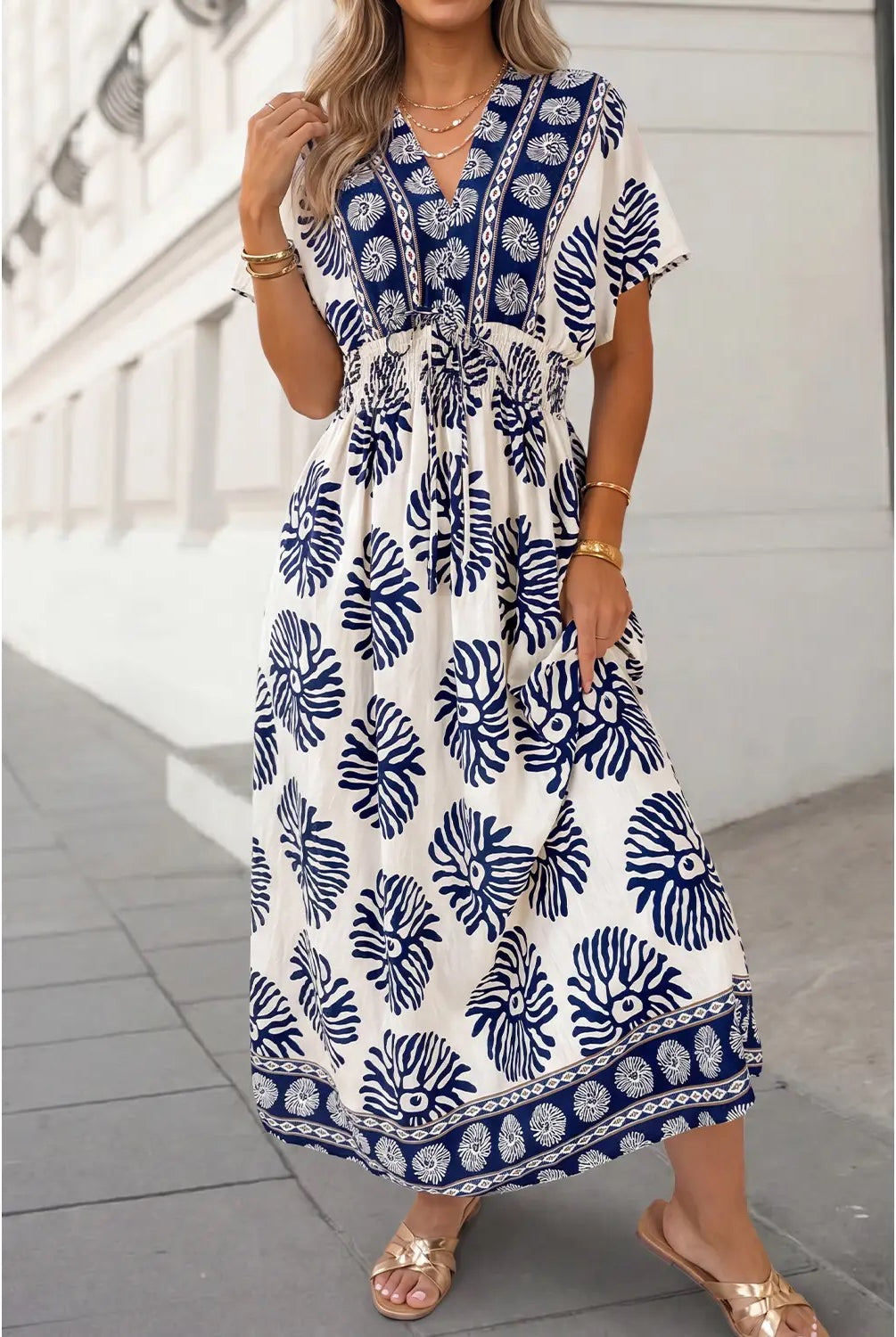 A woman wearing a white maxi dress with a navy blue tropical leaf and border print, standing outdoors on a paved walkway.