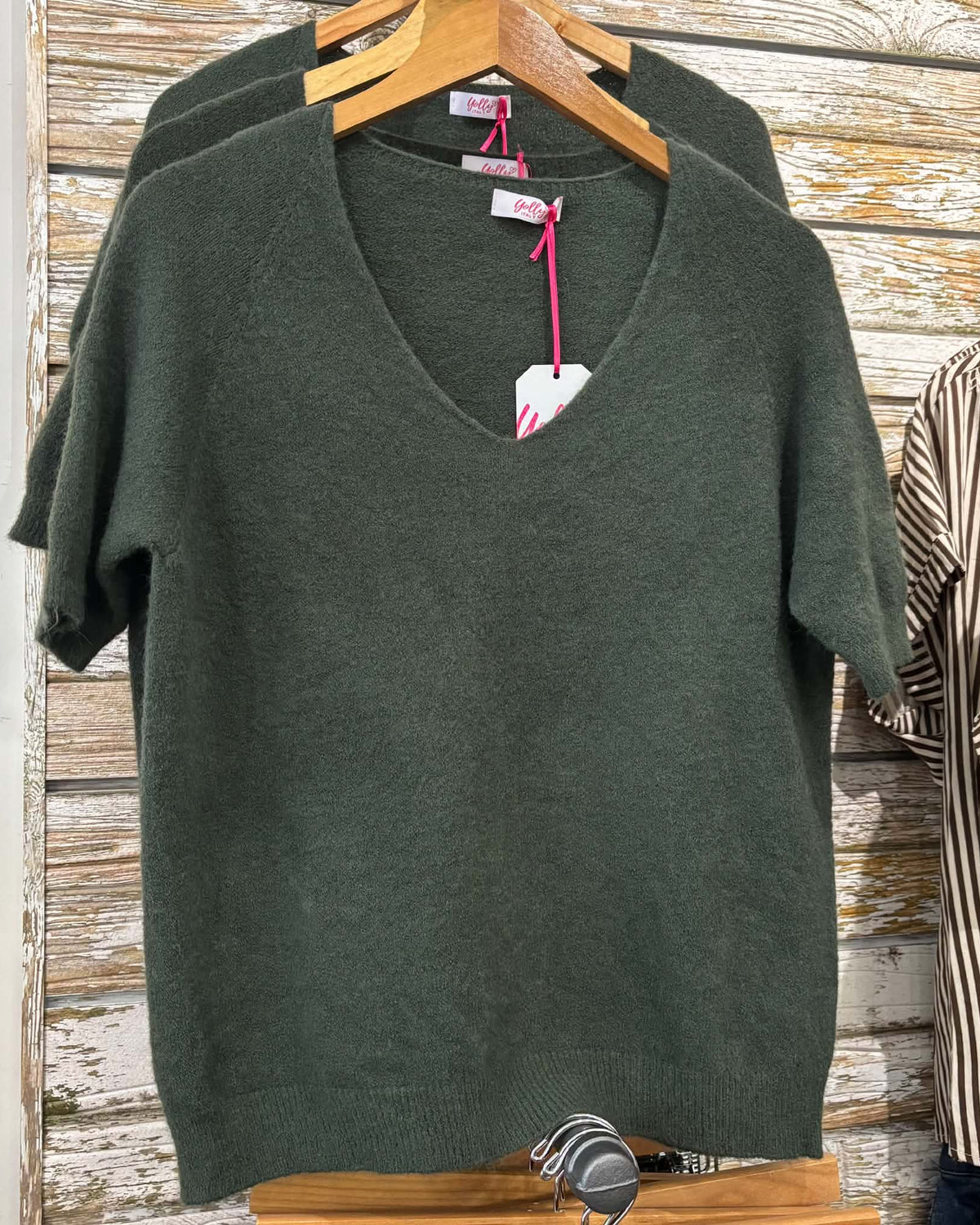 A dark olive green short-sleeve V-neck knit sweater hanging on a wooden hanger against a rustic whitewashed wood plank wall, with another matching sweater partially visible behind it and a striped shirt on the right.
