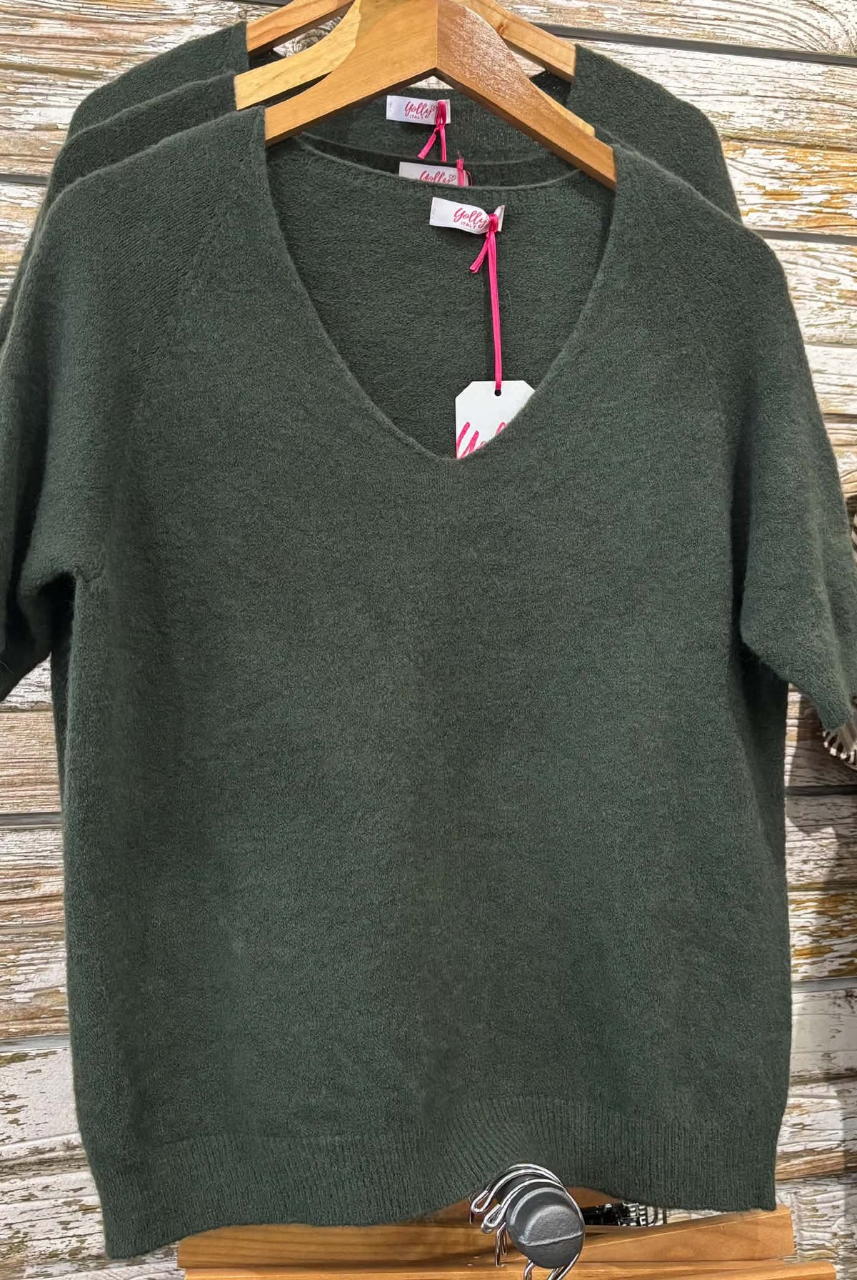 A dark olive green short-sleeve V-neck knit sweater hanging on a wooden hanger against a rustic whitewashed wood plank wall, with another matching sweater partially visible behind it and a striped shirt on the right.