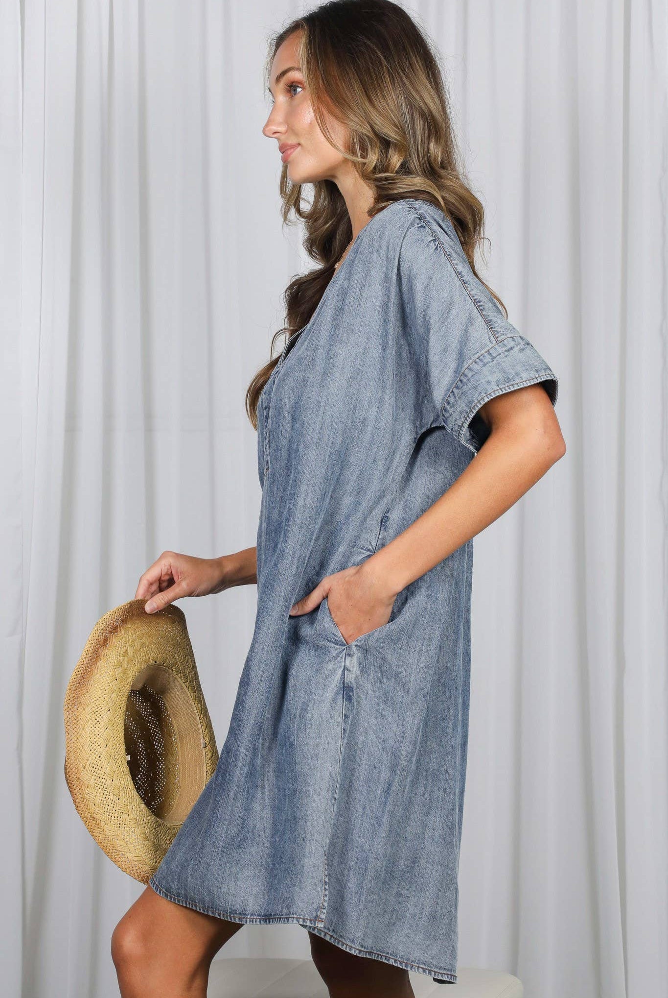 Woman wearing a denim dress holding a hat against a white curtain background