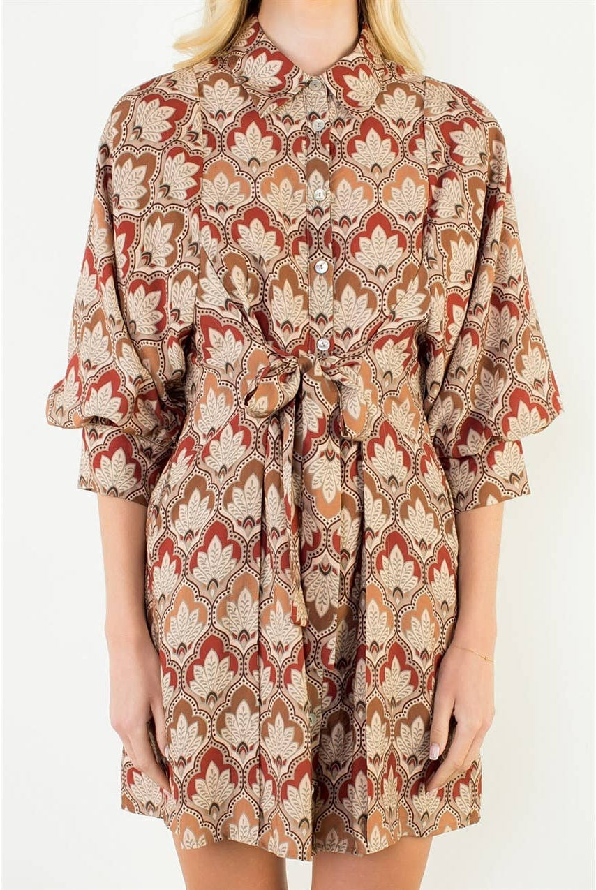 A woman wearing a short collared shirt dress with a beige, rust red, and brown geometric pattern, featuring three-quarter sleeves and a matching waist tie.