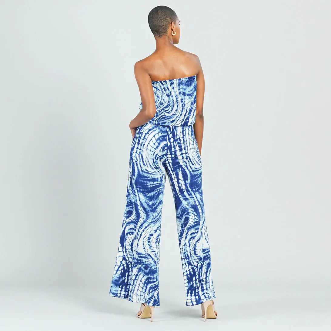 A woman shown from the back wearing a strapless wide-leg jumpsuit with a blue and white tie-dye swirl pattern, paired with beige high heels, against a light gray studio background.