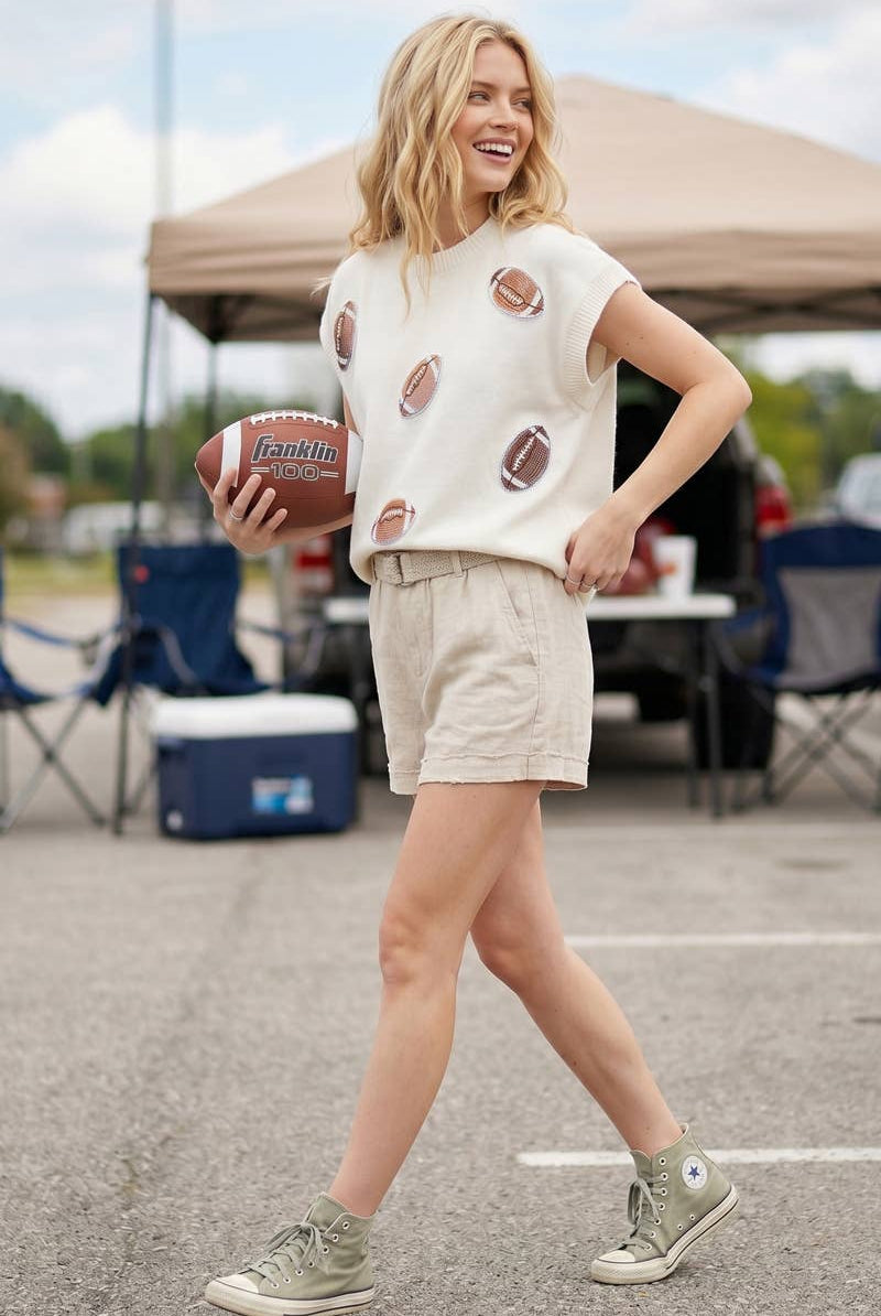 A woman stands outdoors in a parking lot holding a football, wearing a cream short-sleeve knit top with football patches, beige shorts, and green sneakers. A canopy tent and folding chairs are blurred in the background.
