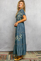 Woman wearing a long denim dress with floral embroidery against a gray wall.