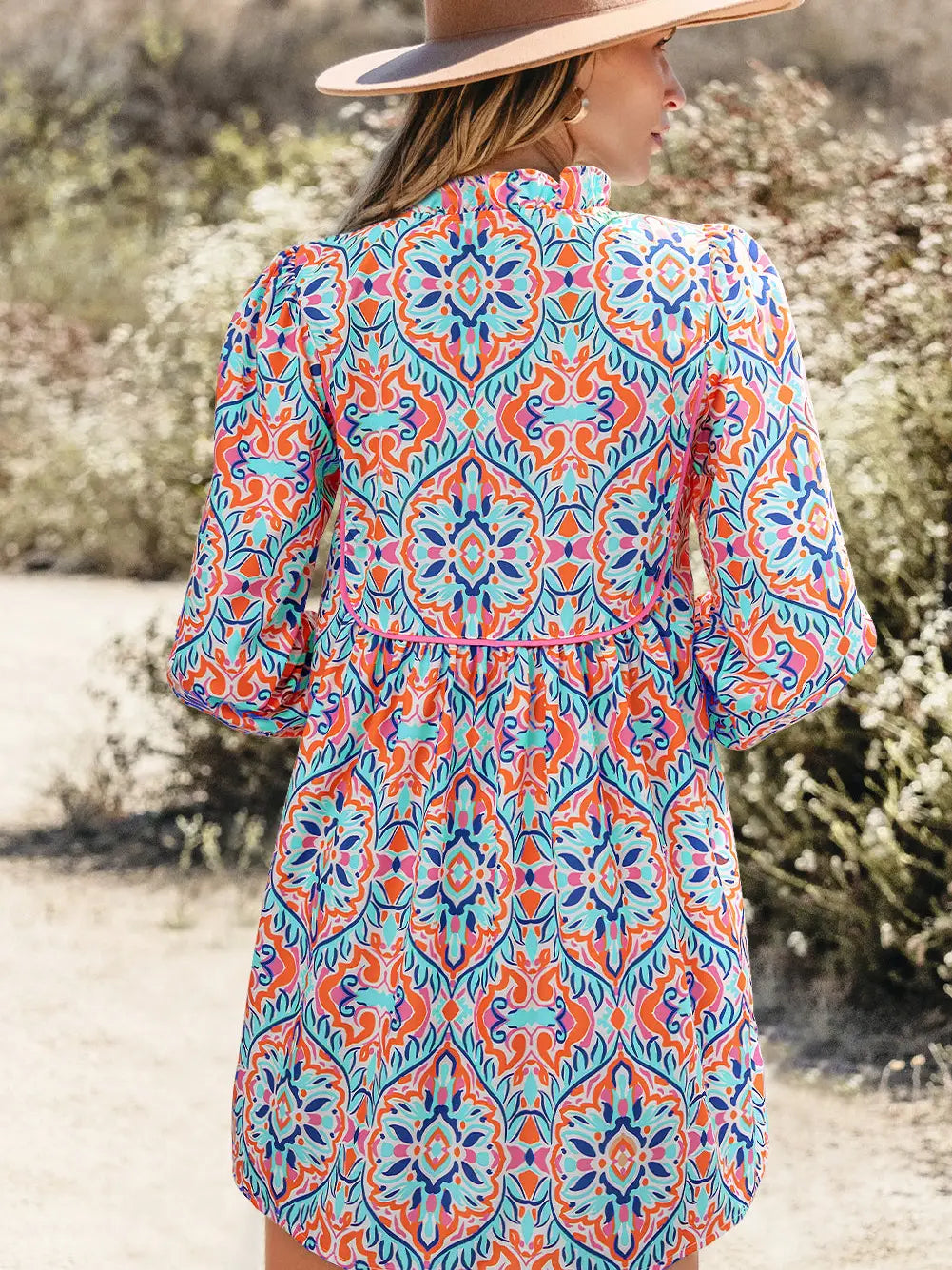 A woman wearing a colorful bohemian-style patterned tunic dress and a wide-brimmed hat, photographed from behind in an outdoor desert setting.