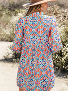 A woman wearing a colorful bohemian-style patterned tunic dress and a wide-brimmed hat, photographed from behind in an outdoor desert setting.