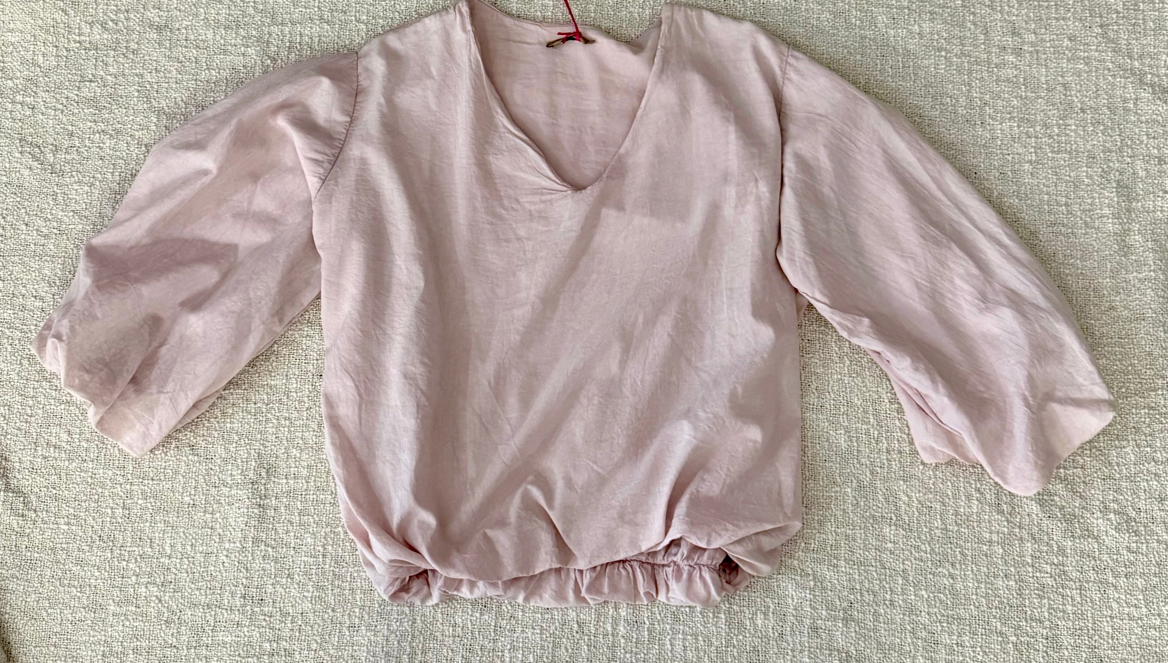 A light pink long-sleeve blouse with a V-neckline and gathered ruffled hem, laid flat on a textured beige fabric background.