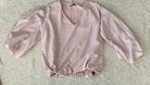 A light pink long-sleeve blouse with a V-neckline and gathered ruffled hem, laid flat on a textured beige fabric background.