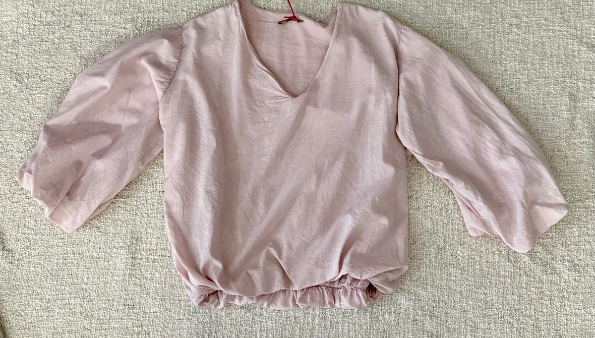 A light pink long-sleeve blouse with a V-neckline and gathered ruffled hem, laid flat on a textured beige fabric background.