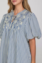 A close-up of a woman wearing a blue and white vertically striped blouse with puff sleeves and a smocked neckline.